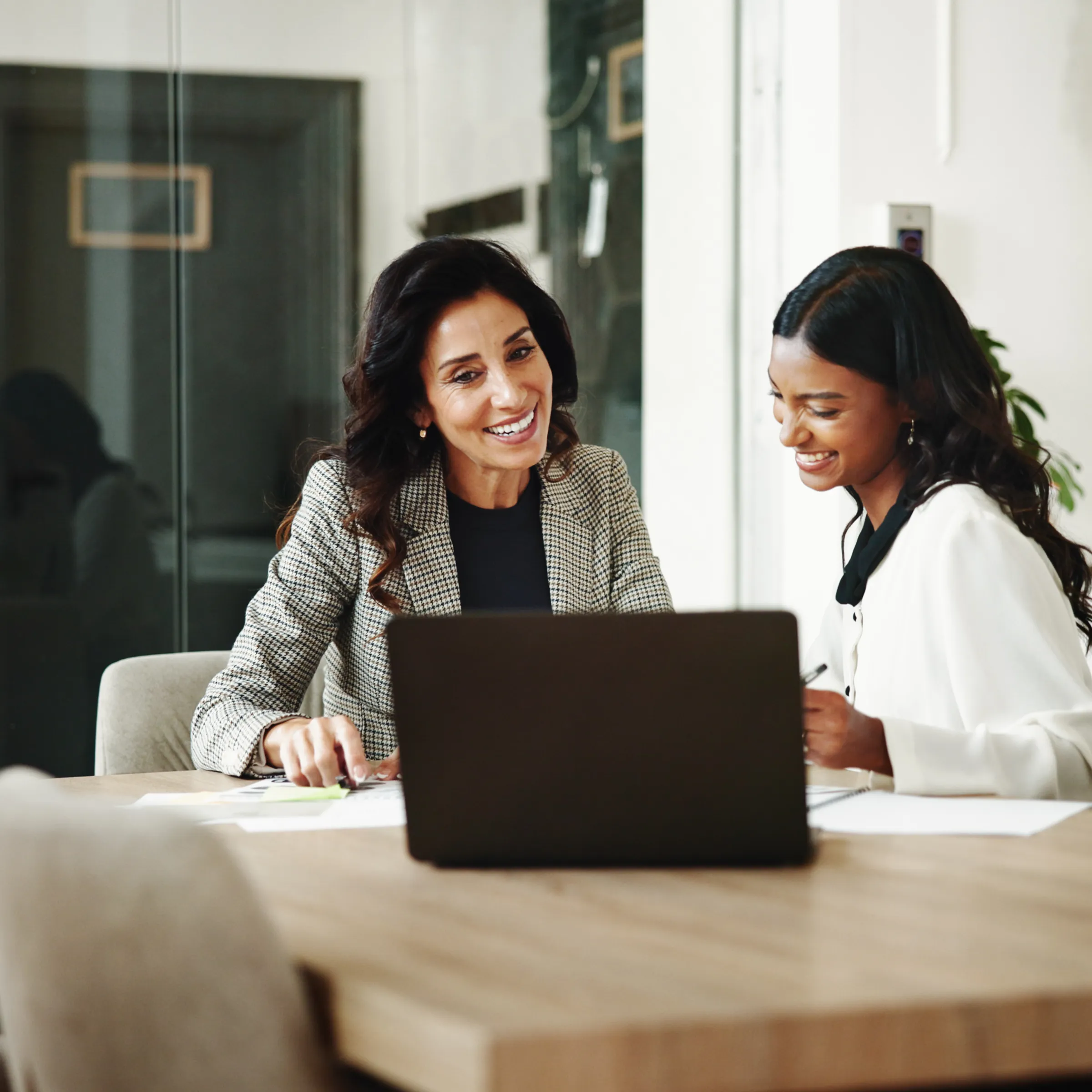 Two smiling women, one in a houndstooth blazer and one in a white blouse, working together at a laptop on a wooden table in a bright office