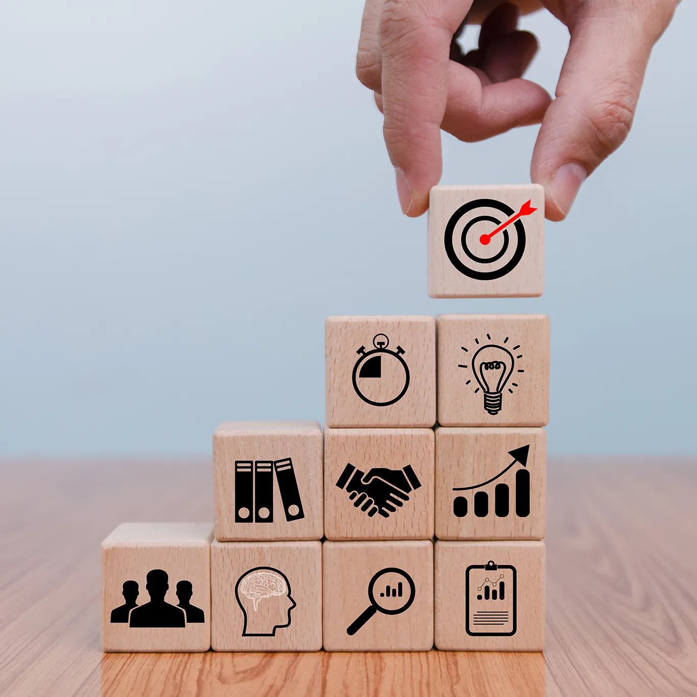 A hand placing a wooden block with a target-and-arrow icon on top of a stepped stack of blocks showing icons for time, ideas, documents, partnership, growth, people, thinking, analysis and reports
