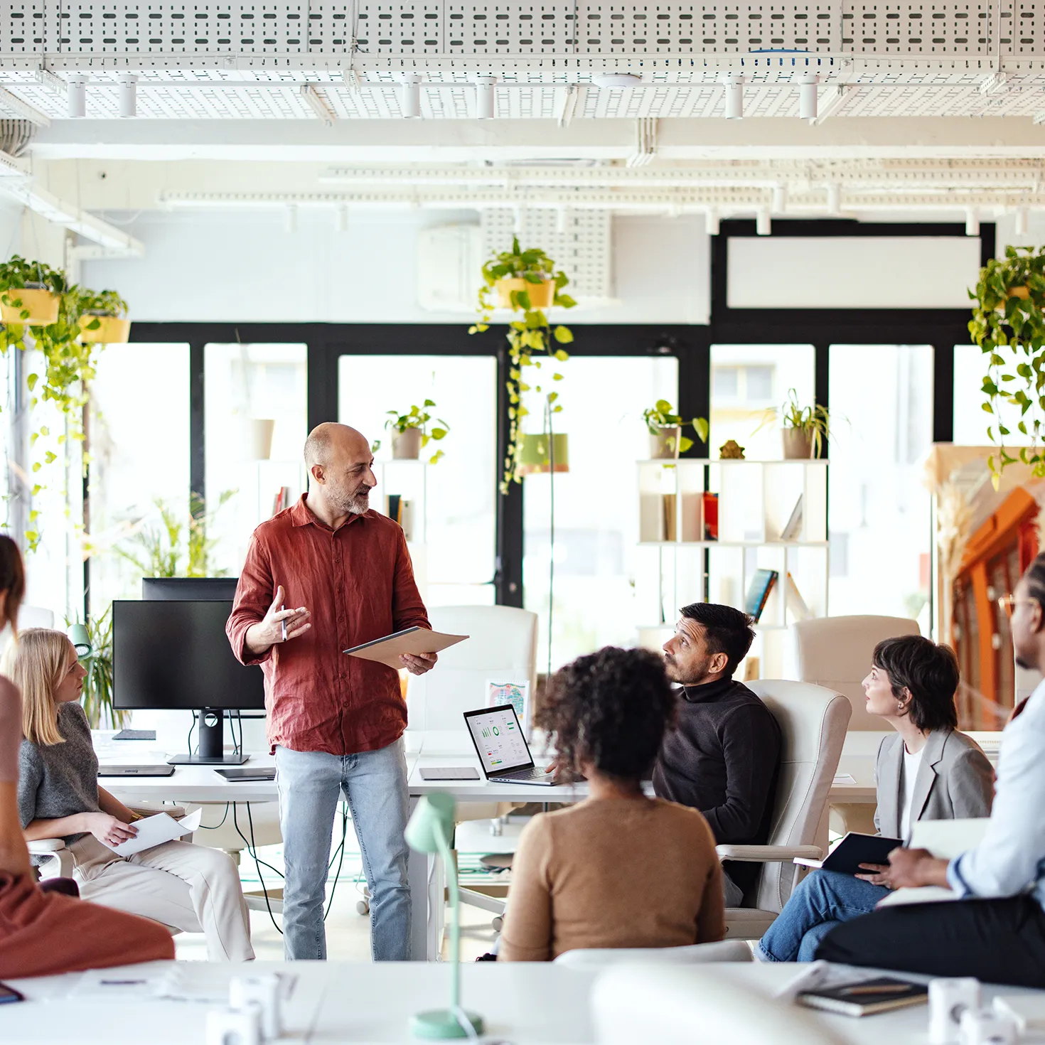 A man in a red shirt holding a tablet leads a discussion with seated colleagues in a bright, plant-filled open office